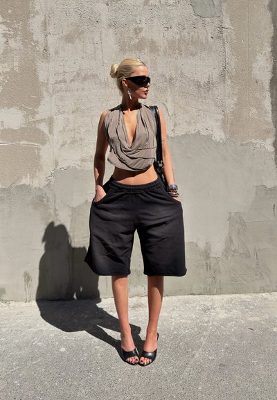 Mujer con shorts negros extragrandes, top sin mangas beige drapeado, gafas de sol negras y tacones negros, de pie contra una pared de hormigón rugosa bajo la luz solar.