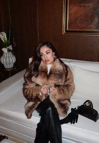 Lussuoso cappotto di pelliccia marrone con ampio colletto, indossato sopra una maglietta bianca, abbinato a pantaloni in pelle nera e una piccola borsa nera.