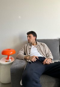 Γκρι καναπές με μπεζ ζακέτα πάνω από λευκό πουκάμισο; πορτοκαλί στρογγυλή λάμπα σε τραπεζάκι πλάγιας από υφή πέτρας; τατουάζ στα χέρια.