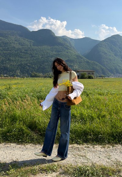 Joven mujer de pie en un camino sosteniendo flores silvestres amarillas en un campo con montañas verdes y cielo azul al fondo.