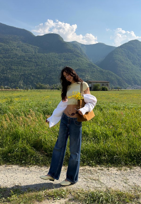Mladá žena stojí na cestě a drží žluté divoké květiny na poli s zelenými horami a modrou oblohou v pozadí.