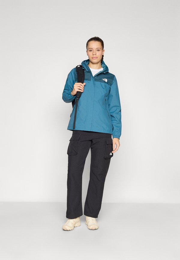 Jachetă impermeabilă de culoare teal cu glugă, accente negre și buzunare cu fermoar. Asortată cu pantaloni cargo întunecați și pantofi bej, potrivită pentru activități în aer liber.