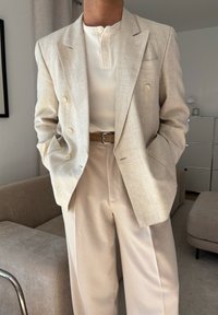 Blazer bege em linho com botões duplos, combinado com uma henley branca com nervuras e calças bege. Tecido texturizado e corte ajustado.