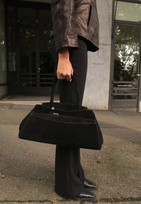 Borsa nera in suede con un design geometrico, forma strutturata, doppio manico e un accento argentato. Indossata da una persona con pantaloni neri.