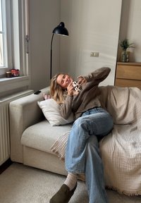 Jasnoszara kanapa z materiału, na której leży teksturowany koc i poduszki. Osoba siedzi, trzymając wzorzystą filiżankę, ubrana w brązowy sweter i niebieskie dżinsy.