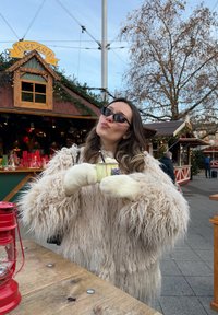 Vêtement d'extérieur beige duveteux, gants blancs, tenant une tasse jaune clair avec un dessin. Lanterne sur une table en bois et étals festifs en arrière-plan.