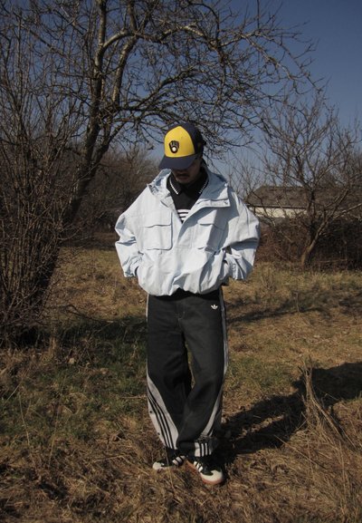 Persona con chaqueta azul claro, pantalones negros de Adidas y una gorra de béisbol amarilla y azul marino, de pie con las manos en los bolsillos al aire libre sobre hierba seca.