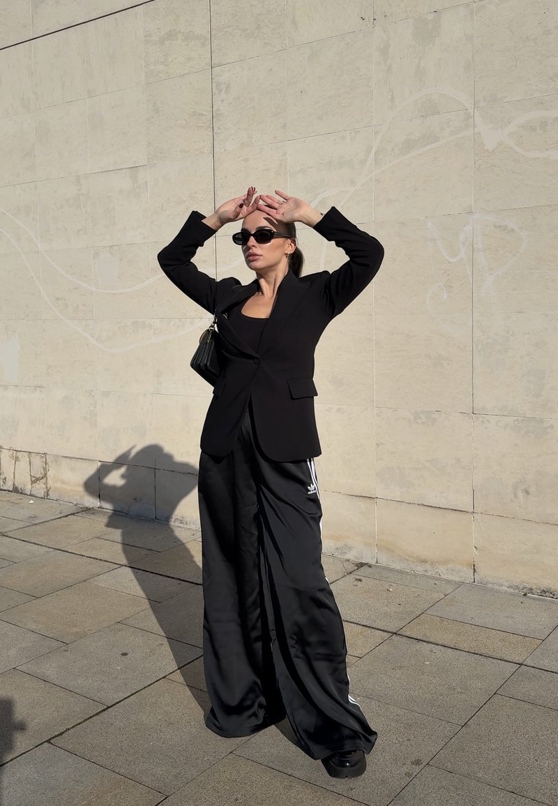 Donna con blazer nero, pantaloni larghi da tuta neri, occhiali da sole e borsa a tracolla posa contro un muro piastrellato beige sotto la luce del sole.