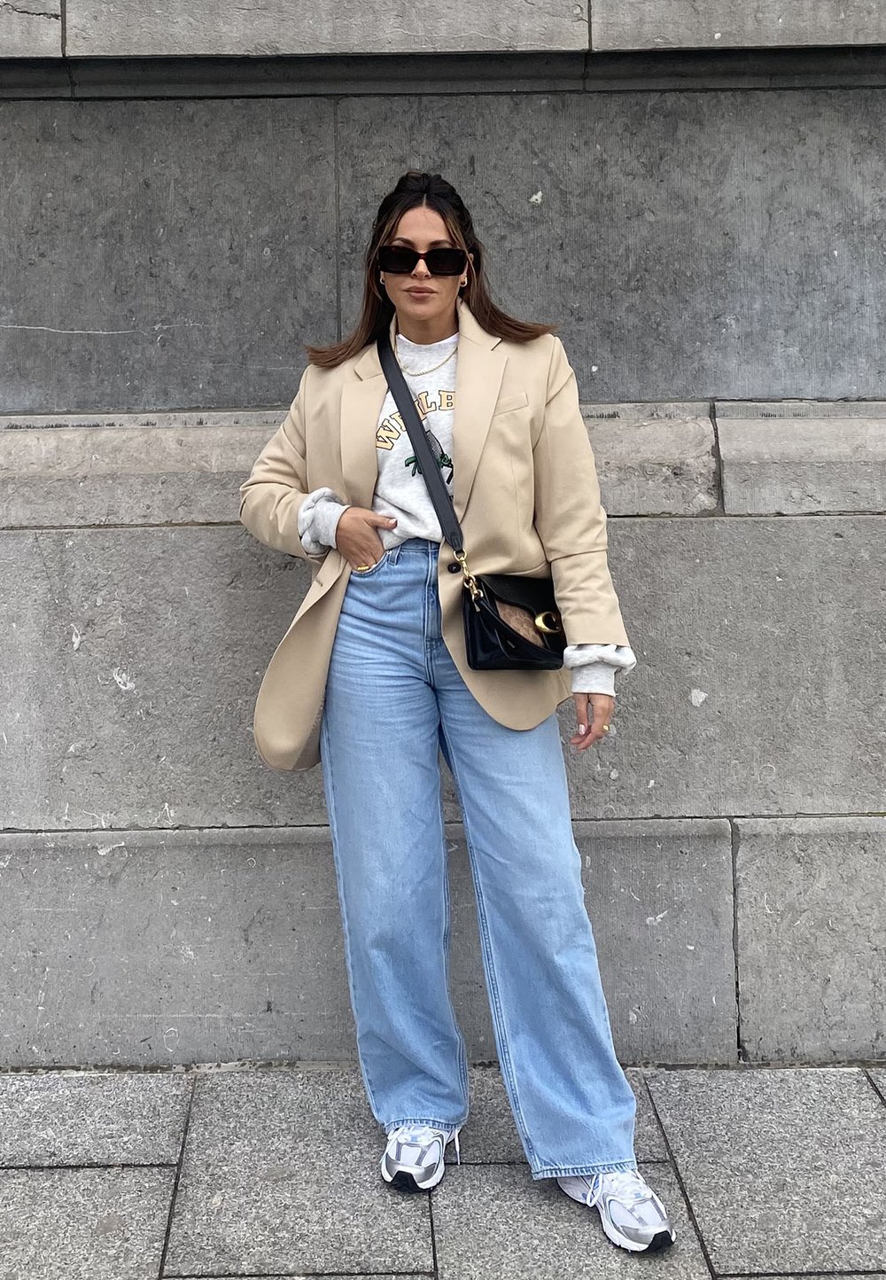 Blazer oversize beige, sudadera gris con texto verde, jeans de pierna ancha azul claro y zapatillas blancas. Bolso negro cruzado, gafas de sol.