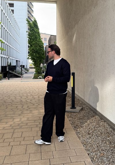 Suéter azul marino sobre camisa blanca, pantalones negros y zapatillas plateadas. De pie sobre un camino de piedra con edificios modernos y vegetación al fondo.