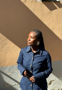 Camicia di denim blu scuro con colletto strutturato, bottoni in ottone e maniche lunghe. Modello che tiene occhiali, posato su una parete texturizzata.
