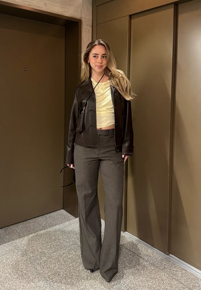 Mujer con cabello largo y rubio, vestida con una chaqueta de cuero negra, camiseta de color amarillo pálido, pantalones grises de pata ancha, sosteniendo gafas de sol en un pasillo con paredes marrones.