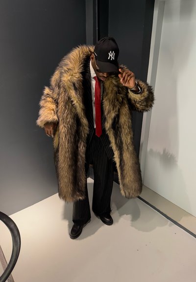 Hombre con un gran abrigo de piel marrón sobre un traje de rayas, corbata roja, zapatos negros y una gorra negra de los New York Yankees ajustándose la gorra.