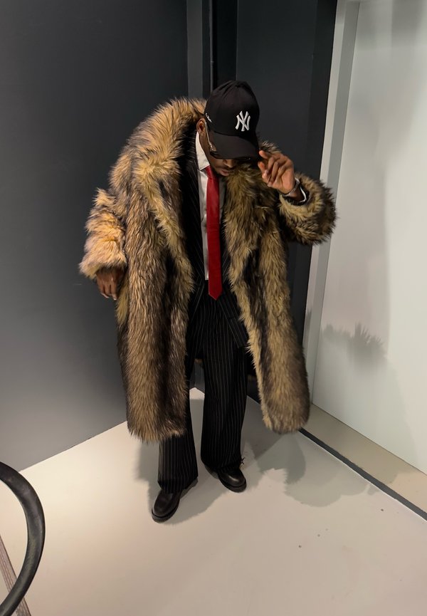 Man draagt een grote bruine bontjas over een streepjespak, rode stropdas, zwarte schoenen en een zwarte New York Yankees-pet en past de pet aan.