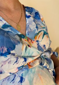 Φλοράλ φόρεμα τύπου περιτύλιξης σε αποχρώσεις μπλε και πορτοκαλί, φτιαγμένο από ελαφρύ ύφασμα. Διαθέτει ζωνάκι στη μέση και βαθύ V ντεκολτέ. Χρυσό κρεμαστό κολιέ.