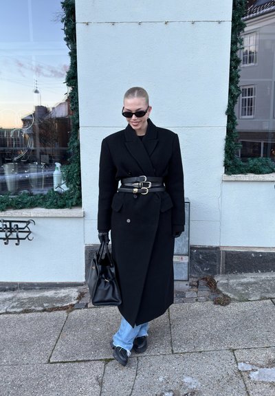 Persona con un largo abrigo negro con tres cinturones, guantes negros, gafas de sol, sosteniendo un bolso negro, de pie en una acera cerca de un edificio.