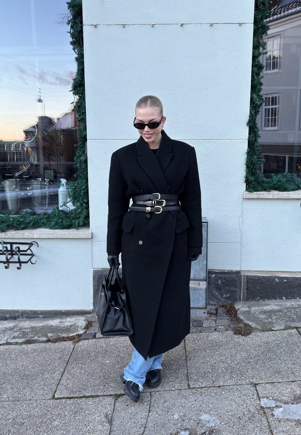 Person wearing a long black coat with three belts, black gloves, sunglasses, holding a black handbag, standing on a pavement near a building.