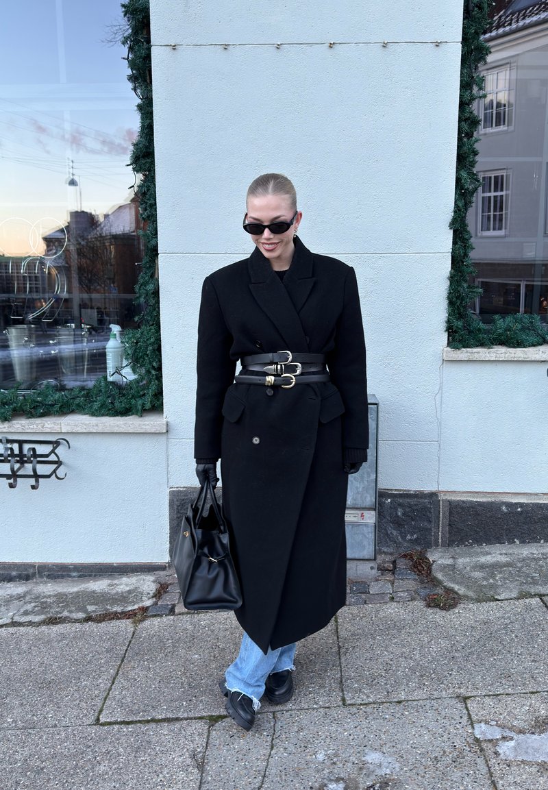 Personne portant un long manteau noir avec trois ceintures, des gants noirs, des lunettes de soleil, tenant un sac à main noir, debout sur un trottoir près d'un bâtiment.