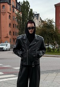 Schwarze Lederbikerjacke mit Reißverschluss- und Druckknopftaschen, getragen über einem schwarzen Hoodie. Das Model trägt dunkle Sonnenbrillen und steht in einer städtischen Umgebung.