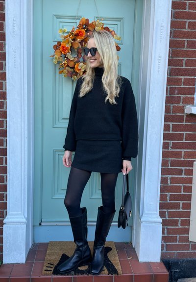Mujer con cabello rubio largo, vestida con un suéter negro, falda, medias y botas, sonríe junto a una puerta azul decorada con una corona de otoño, sosteniendo un bolso negro.