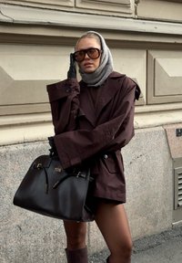 Bruine oversized jas met gerimpelde mouwen, grijze sjaal, zwarte handschoenen, grote donkere handtas en bijpassende bruine kniehoge laarzen.