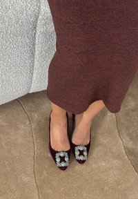Pieds chaussés de hauts talons en velours bordeaux avec de grandes boucles carrées en strass, posés sur un tapis brun clair à côté d'un mobilier en tissu gris.