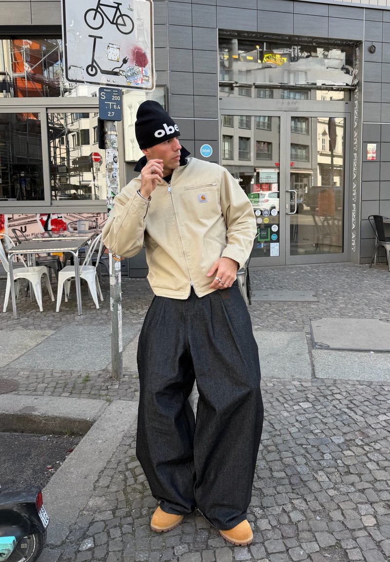 Uomo con giacca beige, berretto nero, pantaloni scuri oversize e stivali color tabacco che posa su un marciapiede in pietra vicino a un cartello per biciclette e scooter.
