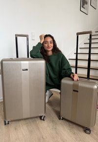 Jeune femme aux cheveux bouclés foncés, vêtue d'un sweat-shirt vert, posant à l'intérieur avec deux valises à roulettes métalliques beige sur un sol en bois.