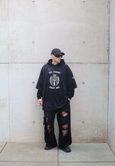 Sudadera negra de gran tamaño con estampado gráfico, combinada con pantalones anchos rasgados negros. Detalles adicionales incluyen una gorra de béisbol y gafas de sol oscuras.