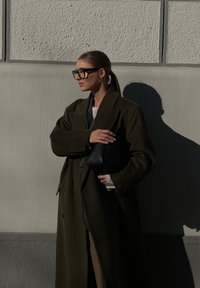 Ελαιογ绿色 μάλλινο παλτό με μεγάλα κουμπιά, φορεμένο πάνω από μαύρο σύνολο, κρατώντας μια μικρή μαύρη υφασμάτινη τσάντα ενάντια σε έναν ανοιχτό γκρι τοίχο.