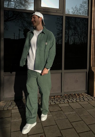 Camisa de pana verde de botones sobre una camiseta blanca, combinada con pantalones verdes holgados y zapatillas blancas, con un fondo de panel de vidrio.