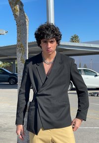 Blazer preto com um ajuste personalizado e um fecho diagonal, usado sobre uma blusa transparente, combinado com calças de cor clara. Os acessórios incluem colares em camadas.