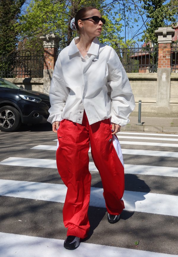 Mujer con chaqueta blanca y pantalones rojos cruzando un paso de peatones con árboles y una cerca de ladrillos en el fondo.