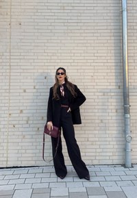 Femme en costume noir et écharpe se tenant contre un mur de briques claires, tenant un sac à main bordeaux, portant des lunettes de soleil et des chaussures pointues.