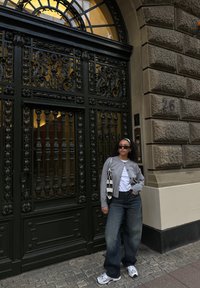 Tenue décontractée avec un cardigan gris, un t-shirt blanc, un jean large foncé et des baskets argentées. Debout à côté d'un portail en fer forgé orné.