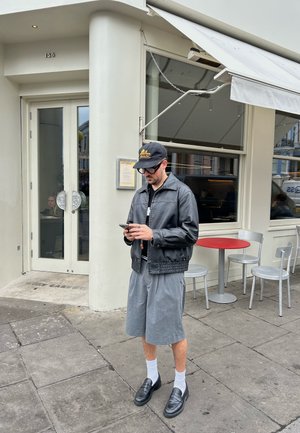 Chaqueta de cuero negra, pantalones cortos grises, calcetines blancos, mocasines negros. De pie frente a un café con grandes puertas de cristal y una mesa roja.