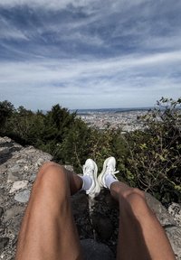 Hvite sportsko med snøring, brukt med hvite sokker, på en steinete avsats som ser ut over en by- og grøntområde under en blå himmel.