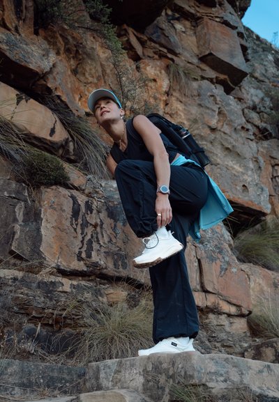 Mujer con ropa de senderismo negra y azul atándose una zapatilla blanca en un sendero rocoso, con mochila y gorra frente a una pared de roca marrón.