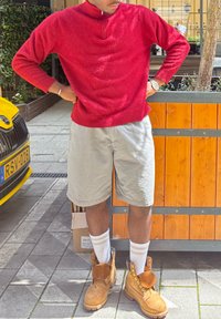 Suéter texturizado vermelho com gola zíper, calções cinzentos, meias brancas com riscas e botas de trabalho bege num passeio de ladrilhos perto de elementos em madeira e amarelos.