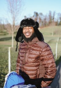 Doudoune marron avec un logo Champion, accompagnée d'un chapeau noir en fausse fourrure. En arrière-plan, un paysage herbeux avec des arbres.