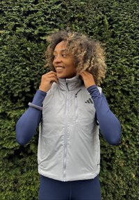 Viegls sudraba veste ar augstu apkakli, ar rāvējslēdzēju un logotipu. Uzvilkta virs tumši zilas garām piedurknēm blūzes, fons ir zaļš dzīvžogs.