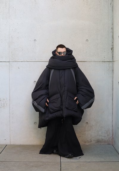 Abrigo negro acolchado de gran tamaño con mangas amplias, cuello alto y acentos grises; combinado con pantalones negros de pierna ancha y gafas de sol.