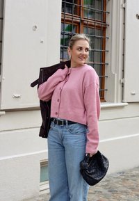 Růžový pletený cardigan s kulatým výstřihem a knoflíky, spárovaný se světle modrými džínami s širokými nohavicemi a černou tkanou kabelkou.