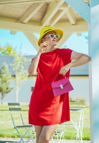 Femme en robe rouge et chapeau jaune tenant un sac à main rose, debout sous un pavillon en bois avec des chaises et des tables d'extérieur en arrière-plan.