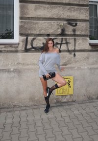 Šedé oversize svetr šaty, černé hodinky, černé tenisky a ponožky po kolena. Betonová zeď s grafity a žlutý elektrický box na pozadí.