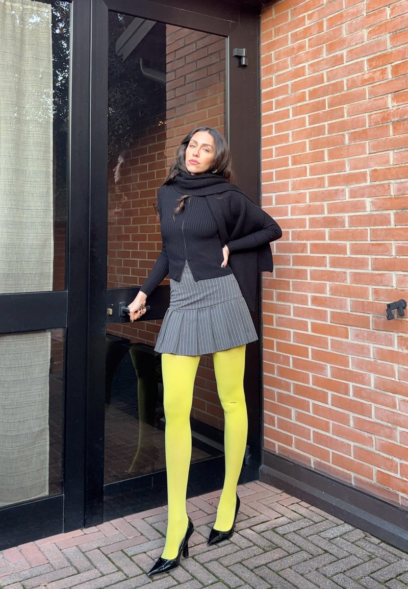 Donna con maglione nero, gonna grigia a righe sottili e collant giallo brillante, in piedi davanti a una porta di vetro con sfondo di muro di mattoni.