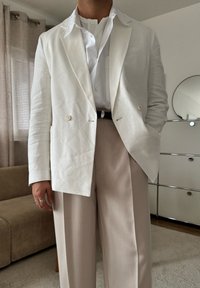 Személy törtfehér, kétsoros gombolású blézert visel fehér gombos ing felett, bézs, magasderekú, pliszírozott nadrágban, beltéren áll.