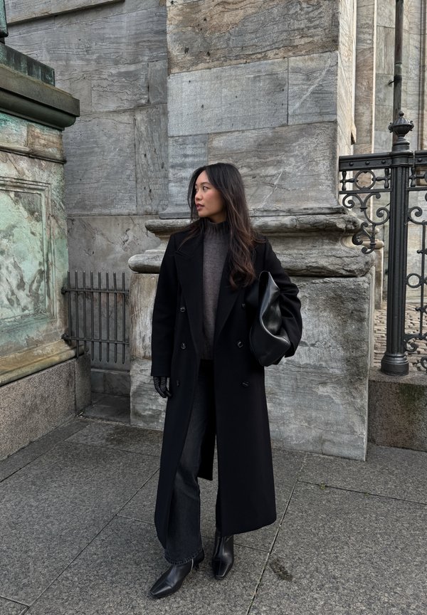 Cappotto nero doppiopetto sopra un maglione grigio, abbinato a jeans neri a gamba larga e stivali alla caviglia con punta. Borsa nera in mano.