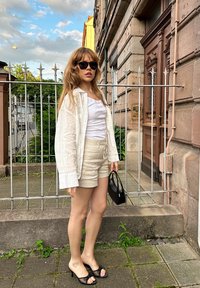 Camicia di lino bianca sopra una canotta bianca, pantaloni corti beige con cordoncino, borsa nera e sandali con tacco neri. Sullo sfondo si vedono un edificio e vegetazione.