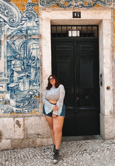 Puerta doble negra con una reja de metal decorativa, situada frente a intrincados azulejos azules y blancos que presentan imágenes históricas. Persona joven con una camiseta clara y pantalones cortos de mezclilla se encuentra al lado.